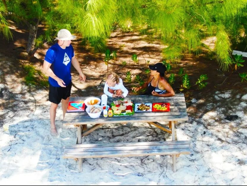 A family picnic laid out on a wooden table on the sand, in the shade of the trees