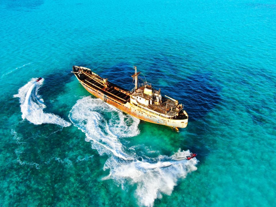 Two jet skis carving wakes around a rusting shipwreck in turquoise water, seen from above